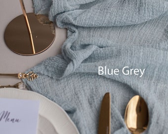 May include: Blue grey textured fabric runner with gold cutlery and a white plate with a menu card on top. The fabric is folded and draped over a table.