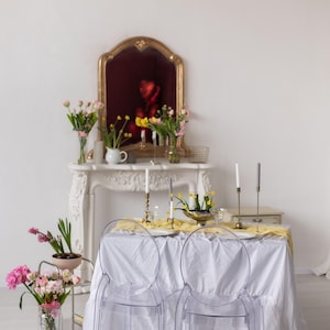 May include: A white tablecloth with a yellow runner covers a table set for two with clear ghost chairs. The table is decorated with candles, flowers, and a vase of yellow flowers. A white marble fireplace mantel with a gold mirror hangs above the table.