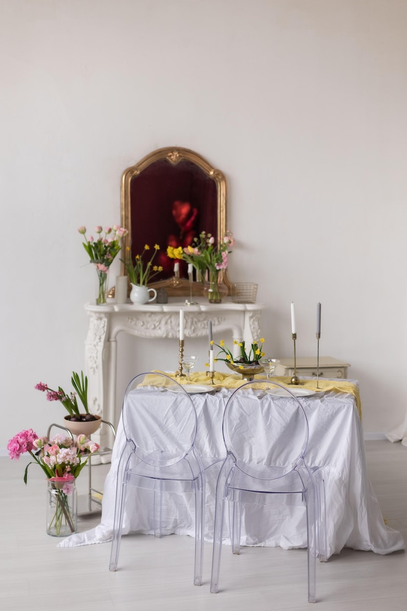 May include: A white tablecloth with a yellow runner covers a table set for two with clear ghost chairs. The table is decorated with candles, flowers, and a vase of yellow flowers. A white marble fireplace mantel with a gold mirror hangs above the table.