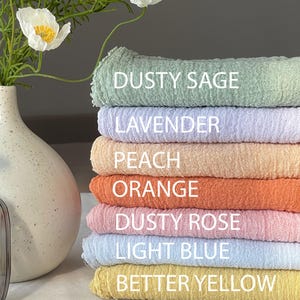 Op de afbeelding: Een stapel van zeven stoffen vierkanten in verschillende pastelkleuren, waaronder stoffig salie, lavendel, perzik, oranje, stoffig roze, lichtblauw en beter geel. Elk vierkant heeft een andere kleur en is gelabeld met de naam van de kleur.