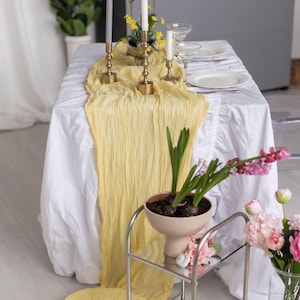 May include: A yellow table runner with a ruffled edge is draped over a white tablecloth.  The runner is made of a soft, sheer fabric and has a delicate, flowing texture.  A set of candlesticks with white candles are placed on the runner, along with a small potted plant.  The table is set for a dinner party, with plates, silverware, and glasses.