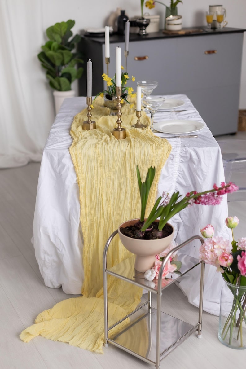 May include: A yellow table runner with a ruffled edge is draped over a white tablecloth.  The runner is made of a soft, sheer fabric and has a delicate, flowing texture.  A set of candlesticks with white candles are placed on the runner, along with a small potted plant.  The table is set for a dinner party, with plates, silverware, and glasses.