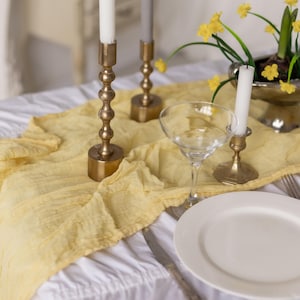 May include: A white tablecloth with a yellow linen runner, three gold candle holders with white candles, a clear glass goblet, and a white dinner plate.