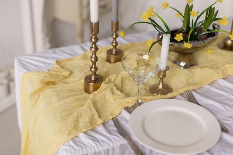 May include: A white tablecloth with a yellow linen runner, three gold candle holders with white candles, a clear glass goblet, and a white dinner plate.