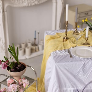May include: A white tablecloth with a yellow runner and a white plate, a glass, and two candlesticks with white candles on a table setting. There are flowers in a vase on a side table.