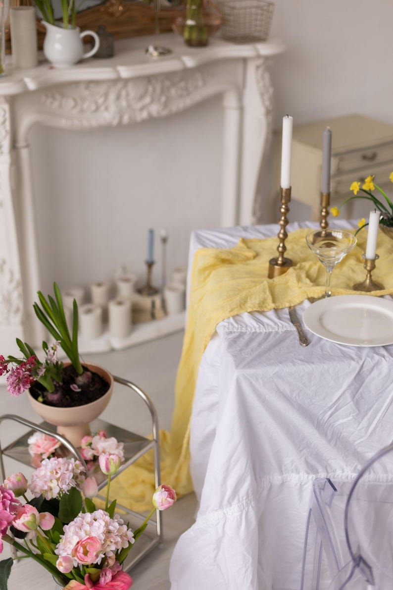 May include: A white tablecloth with a yellow runner and a white plate, a glass, and two candlesticks with white candles on a table setting. There are flowers in a vase on a side table.