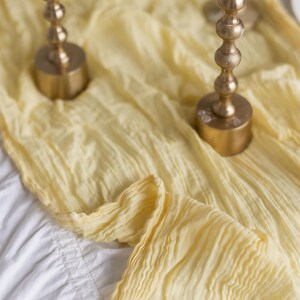 May include: A yellow fabric table runner with two gold candle holders and white candles. The candles are lit and the runner is draped over a white tablecloth.
