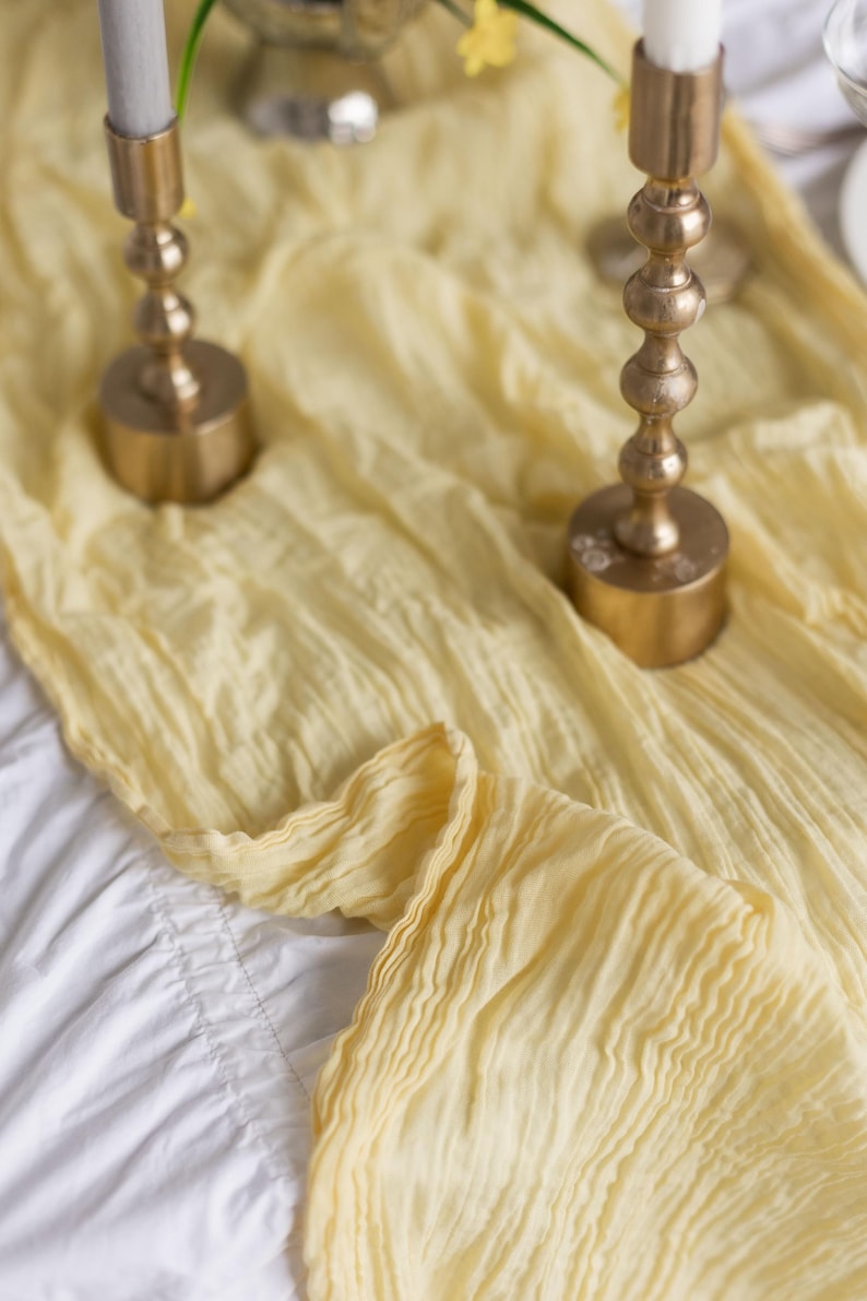 May include: A yellow fabric table runner with two gold candle holders and white candles. The candles are lit and the runner is draped over a white tablecloth.