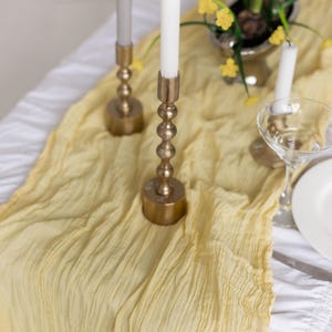 May include: A yellow table runner with a crinkled texture, two gold candle holders with white candles, and a bouquet of yellow flowers in a silver vase.