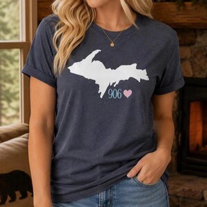 Peut inclure: Un t-shirt à manches courtes bleu chiné foncé arborant un motif blanc représentant la péninsule supérieure du Michigan. Sous la carte, le texte 906 est imprimé en blanc, suivi d'une petite icône de cœur rose clair sur la poitrine.