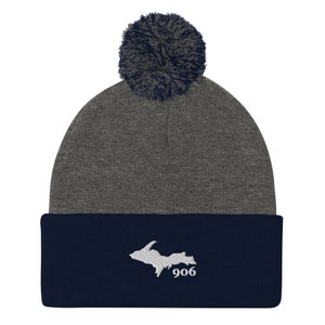 Può includere: Un berretto grigio e blu navy con un pompon grigio scuro. Il polsino blu navy presenta un contorno bianco ricamato della Penisola Superiore del Michigan e il numero "906".
