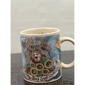 May include: Cream-colored ceramic mug featuring a teddy bear wearing a sunflower hat and holding sunflowers. The mug has the text "LOYAL ORDER OF FRIENDS OF BOYDS" and "Sunny Day Sunflowers". The mug has a curved handle and a blue background.