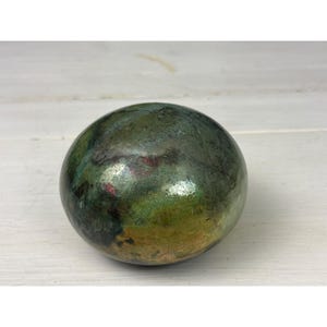 May include: A smooth, spherical stone featuring a mottled surface with deep forest green, olive, and dark charcoal tones, accented by small, subtle flecks of reddish-brown and a polished, reflective finish that catches the light.