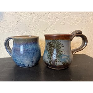 May include: Two ceramic mugs. One is blue with a dark rim and handle. The other mug has a brown rim and handle, with a tree design on the front. The mugs are on a black surface.
