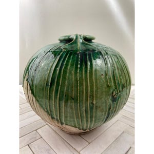 Puede incluir: Un jarrón grande, redondo, de cerámica verde con un diseño texturizado en forma de hoja. El jarrón tiene un esmalte blanco en la parte inferior y un esmalte verde en la parte superior.