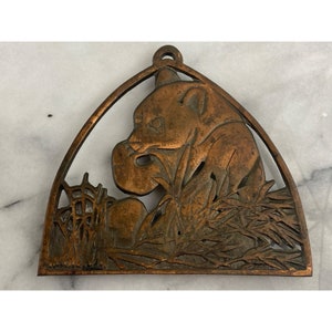 May include: A copper-colored metal wall hanging featuring a panda bear eating. The panda is surrounded by detailed foliage and enclosed within a triangular frame with a loop for hanging. The design has a vintage aesthetic.