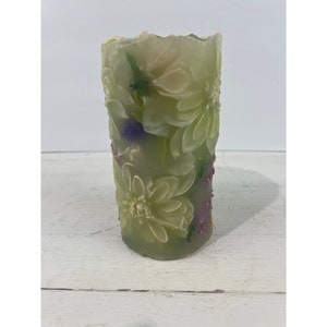 May include: A cylindrical candle with a floral design. The candle is mainly green with white and purple accents. The flowers are sculpted in relief, creating a textured surface. The candle is unscented.