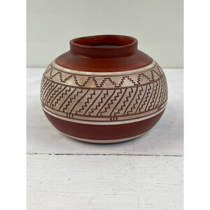 May include: A round, handcrafted ceramic pot with a reddish-brown exterior and a cream-coloured band featuring a geometric pattern. The pot has a small opening at the top and a white base. The design is reminiscent of Native American pottery.