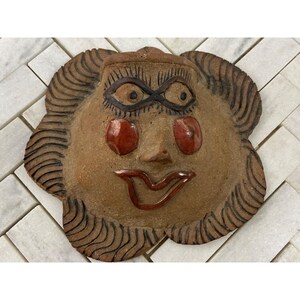 May include: A handcrafted ceramic face mask with a whimsical design. The mask has a brown base colour with red accents on the cheeks and lips. The eyes are intricately detailed, and the hair is stylized with curved lines.