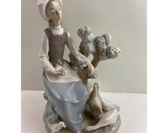 May include: A porcelain figurine depicting a seated figure in a long dress and bonnet, interacting with several geese. The figure is in shades of white, gray, and blue, with a small tree in the background.
