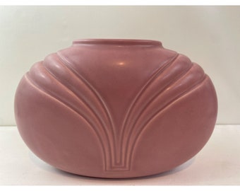 May include: A dusty rose-colored ceramic vase with a rounded, oval shape. The vase features a symmetrical, fan-like design of raised, curved lines on the front. The top has a simple, rounded rim.