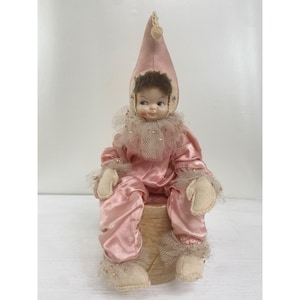 Puede incluir: Muñeca vintage vestida con un disfraz de payaso de satén rosa con un gorro puntiagudo a juego. La muñeca tiene una cara pintada, cabello castaño y está sentada sobre una base cilíndrica. El disfraz presenta adornos de encaje y lentejuelas.