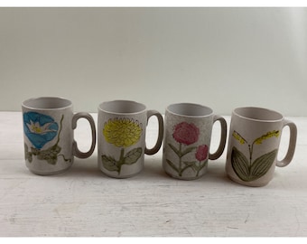 May include: Four ceramic mugs with floral designs. Each mug features a different flower: a blue morning glory, a yellow chrysanthemum, pink flowers, and yellow flowers. The mugs have a neutral beige color with matching handles.