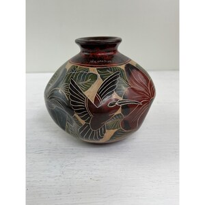 May include: A hand-carved ceramic vase with a dark red rim and a round body. The vase features a hummingbird design with floral and leaf patterns in black, red, and beige. The word "Nicaragua" is written on the vase.