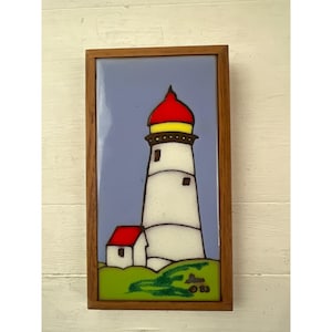 Puede incluir: Azulejo rectangular enmarcado en madera con una escena de faro. El faro es blanco con la parte superior roja y detalles amarillos, sobre un cielo azul claro. Un pequeño edificio con tejado rojo se encuentra en la base, con hierba verde debajo. La obra está firmada y fechada '83.