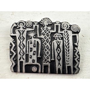 May include: A rectangular, silver-toned metal brooch featuring a stylized design of five figures. The figures have geometric patterns and are set against a black background. The brooch has a textured border and is likely a piece of wearable art.