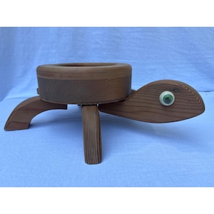 May include: A wooden turtle-shaped planter with a round, recessed top for holding plants. The planter has a light brown finish, with a turtle head and tail. The turtle's eye is a green and white marble. The planter is supported by three legs.