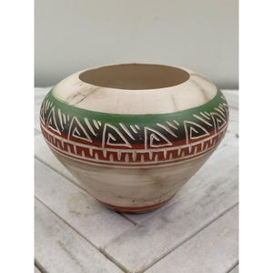 May include: A small, white ceramic pot with a green, red, and brown geometric pattern. The pot is hand-painted and has a slightly rounded shape.