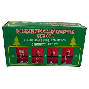 May include: A set of four colourful stocking hangers for Christmas. The hangers are made of high impact plastic and feature Santa, an elf, a snowman, and a drummer boy. The box says "Holiday Stocking Hangers Set of 4".