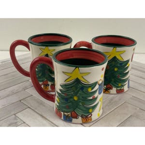 May include: Three ceramic mugs with a Christmas tree design. The mugs are white with green, red, yellow, and blue accents. Each mug has a green Christmas tree with a yellow star on top and red and blue presents underneath. The mugs have red handles and green rims.