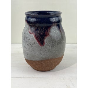 Op de afbeelding: Een handgemaakte keramische vaas met een uniek kleurenschema. De bovenrand is diep marineblauw, overgaand in een gespikkelde grijze body met bordeauxrode druppels. De basis is een natuurlijk, aards bruin. Deze decoratieve vaas is ideaal voor bloemen.