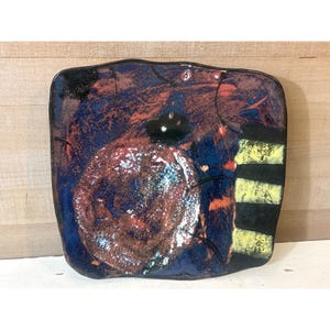 May include: Square ceramic plate with a dark blue background and a large, textured, reddish-brown spiral design. The plate has black and yellow stripes on the right side and a black border. The plate is a unique piece of art.