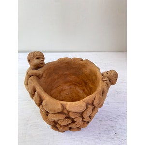 May include: A terracotta-coloured bowl with a textured exterior. Two small figures are sculpted on the sides, appearing to hold the bowl. The bowl's interior is smooth, contrasting with the rough, stone-like exterior. A unique decorative piece.