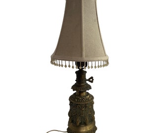 Puede incluir: Una lámpara de mesa antigua con una pantalla de tela beige y una base decorativa de bronce. La lámpara presenta intrincados detalles florales y de volutas, con un fleco de cuentas alrededor del borde inferior de la pantalla. La lámpara mide unos 71 cm de alto.