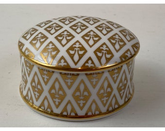 May include: A white and gold decorative trinket box. The box features a repeating pattern of gold fleur-de-lis and diamond shapes. The rim and lid are trimmed with gold. The box is round with a domed lid.