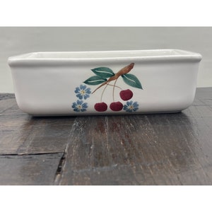 May include: A rectangular white ceramic baking dish with a painted cherry design. The design features three red cherries, green leaves, and blue flowers. The dish is on a dark wooden surface.