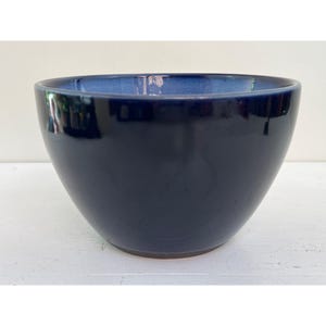 May include: A glossy, navy blue ceramic bowl with a contrasting lighter blue interior. The bowl has a rounded shape and a smooth, reflective surface, suitable for serving or display.