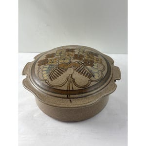 May include: A round, lidded stoneware casserole dish. The lid features a floral design in shades of brown, beige, and blue. The dish has a speckled, earthy tone and side handles. A vintage kitchenware item.