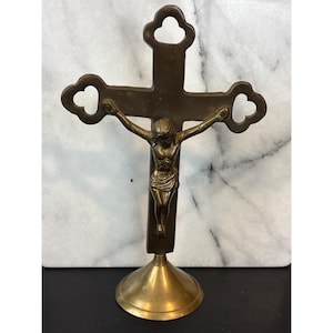 May include: A bronze crucifix with a detailed figure of Jesus Christ. The cross has decorative trefoil ends and is mounted on a round, flared base. The figure is in a crucifixion pose, with arms outstretched.