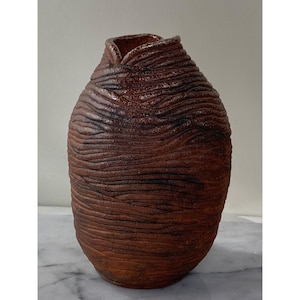 Puede incluir: Un jarrón de cerámica marrón con una superficie texturizada y ondulada. El jarrón tiene una abertura ancha en la parte superior y un fondo redondeado.