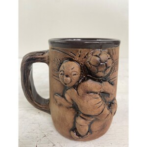 Puede incluir: Taza de cerámica marrón con un diseño esculpido de dos figuras jugando al fútbol. La taza tiene un asa y un borde marrón oscuro, y las figuras están en relieve. La taza es de color marrón medio y parece hecha a mano.