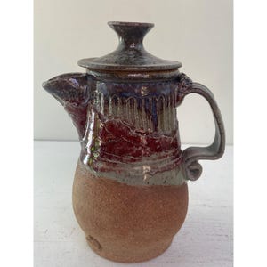 Puede incluir: Una tetera de cerámica hecha a mano con tapa, con un esmalte marrón, rojo y gris. La tetera tiene un cuerpo redondeado y un pico en un lado. El asa es curva y tiene un detalle decorativo en la parte superior.