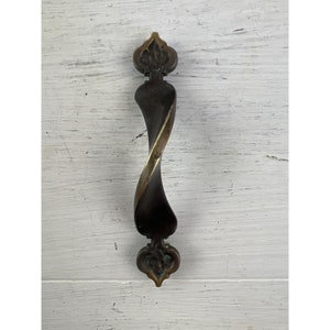 May include: A bronze-toned cabinet pull with a twisted center and decorative ends. The hardware features a dark brown finish with a gold-colored twist in the middle. The ends have a floral design.