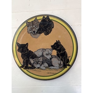 May include: Round decorative plate featuring a wolf pack design. The plate has a tan center with a yellow and green border. The wolves are depicted in black, gray, and tan, with detailed fur textures. The plate is likely ceramic or a similar material.
