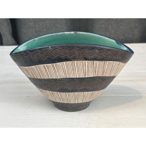 May include: A wide, oval-shaped ceramic bowl with a vibrant turquoise interior. The exterior features a dark, textured surface with alternating horizontal bands of wavy lines and vertical etched stripes in a cream and dark brown color palette.