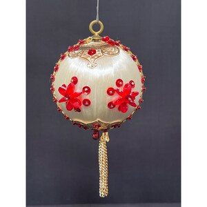Puede incluir: Adorno navideño redondo de color crema con detalles florales rojos y joyas rojas. El adorno tiene una parte superior de metal dorado y una borla de cadena dorada. El adorno cuelga sobre un fondo oscuro.